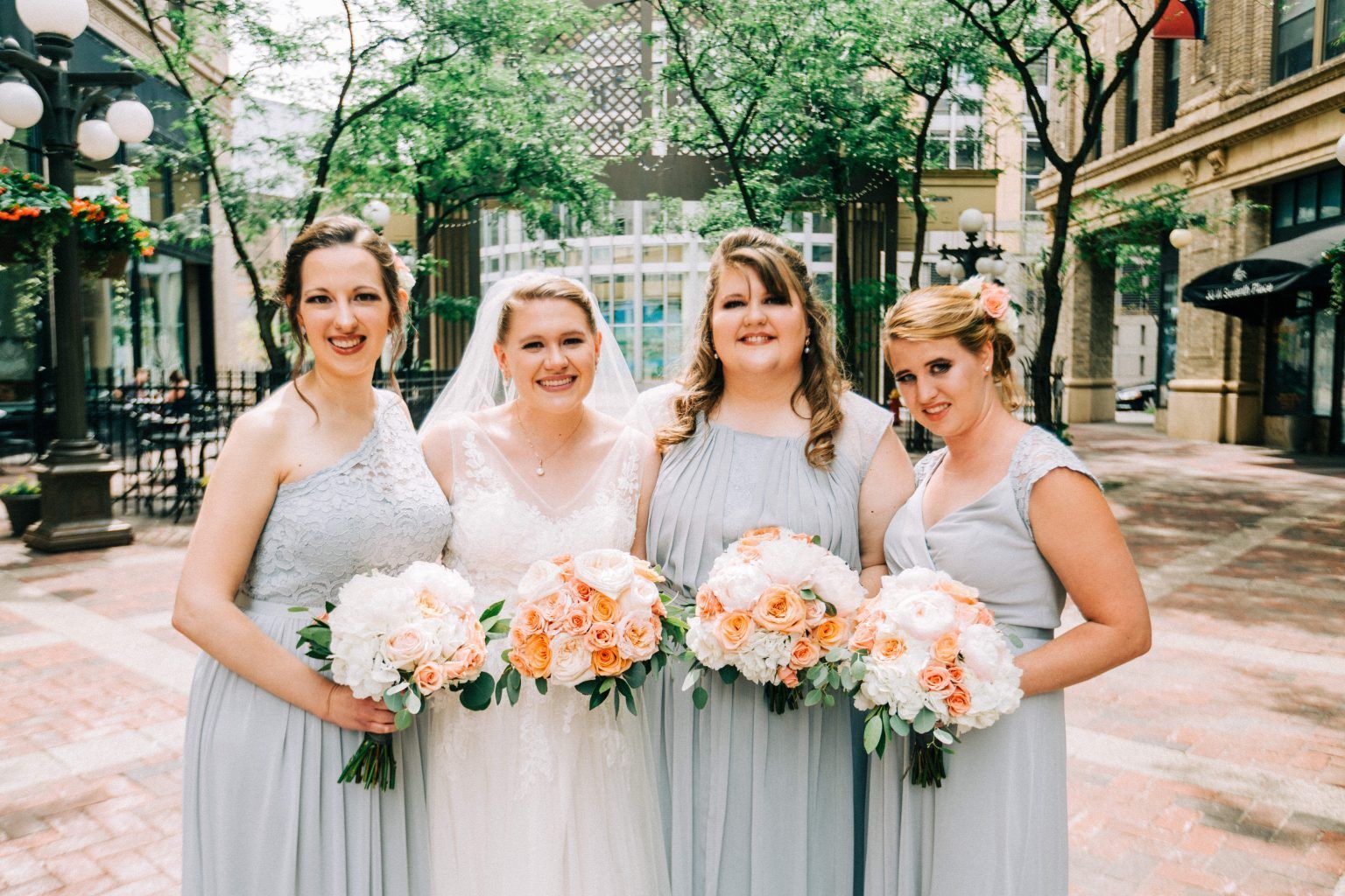 Bride with bridesmaids, light blue dresses, holding peach bouquets, outside, smiling.