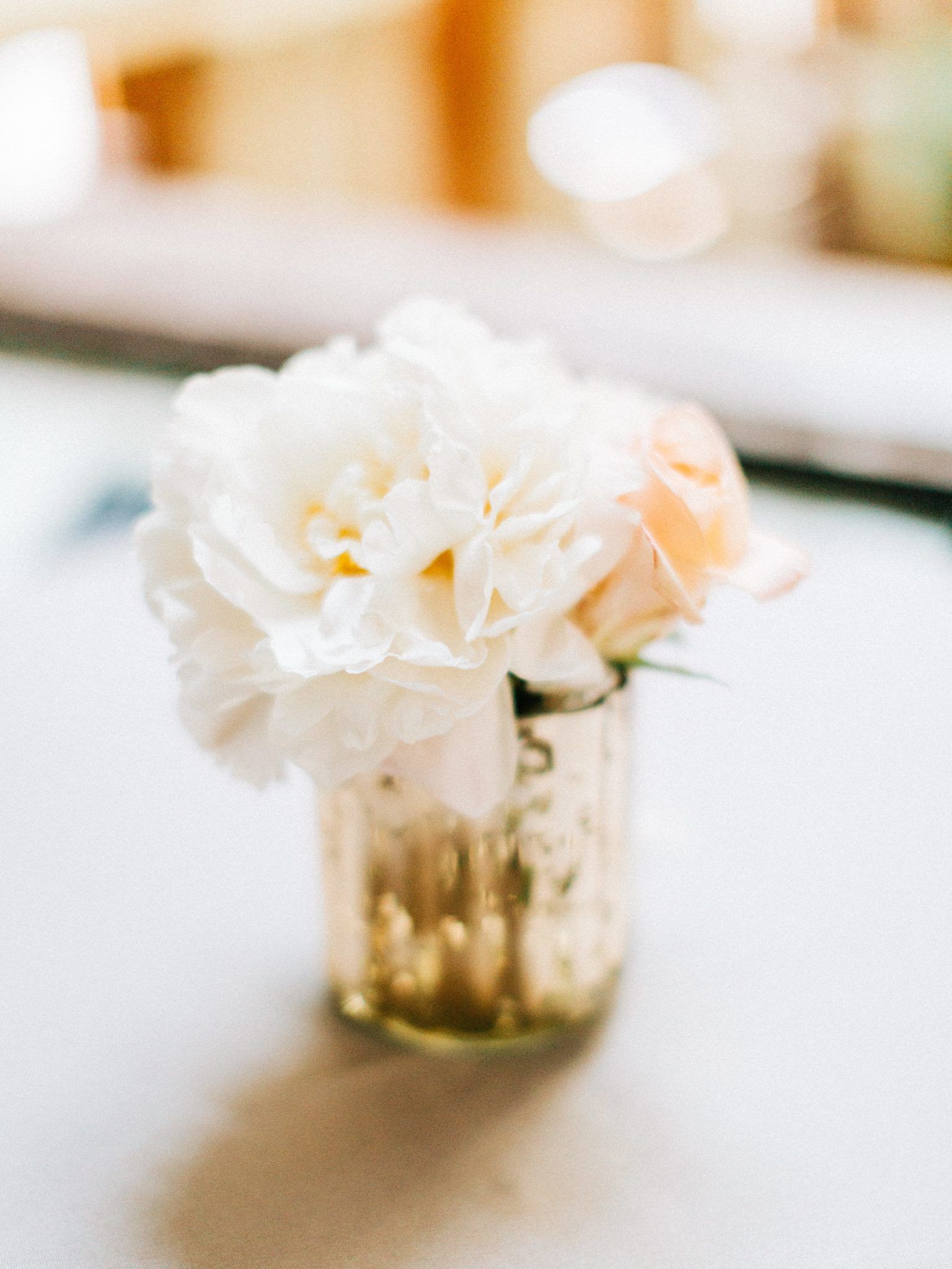 White and peach flowers in a small gold vase on a light-colored surface.