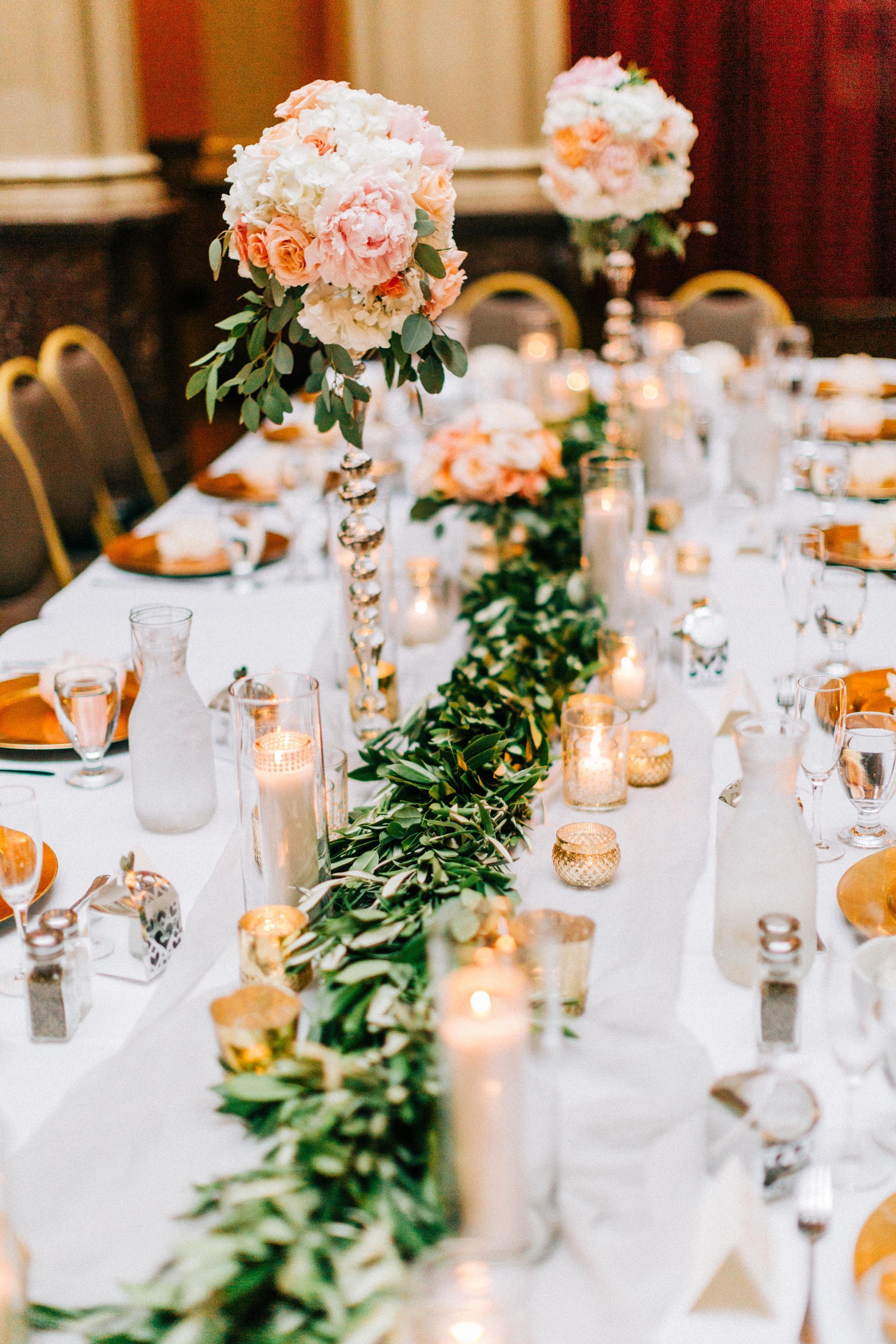 Wedding reception table with floral arrangements, candles, and greenery runner.