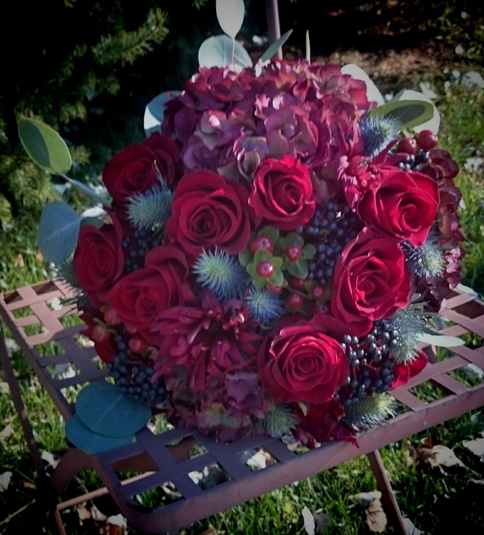 Bouquet of deep red roses, hydrangeas, and thistles on a rusty metal chair in a sunny outdoor setting.