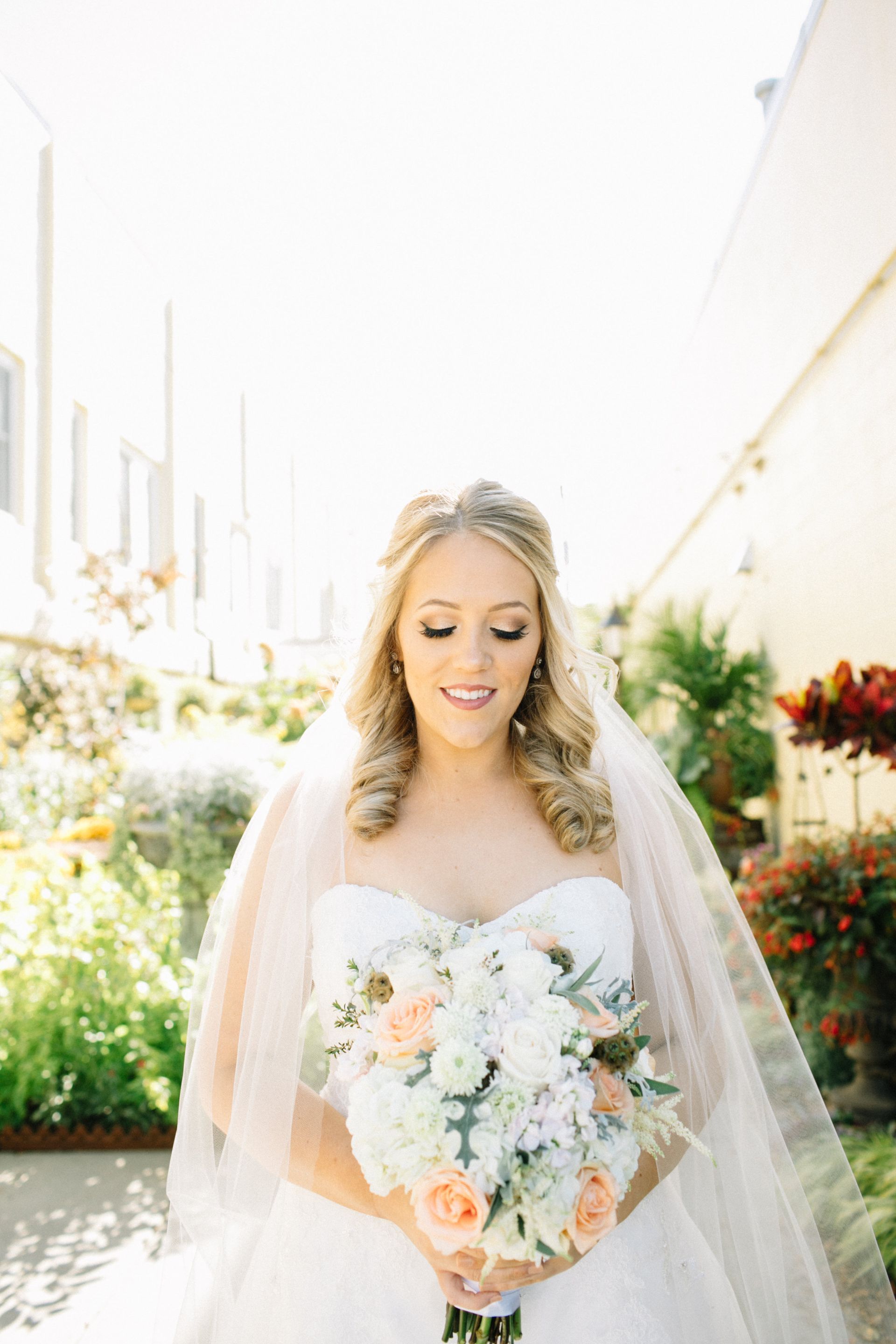 Bride in strapless white dress, veil, holding flowers, looking down, outdoors.