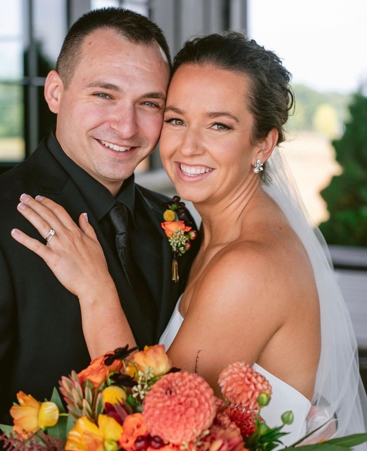 Newlyweds smiling; man in black suit, woman in white dress, holding flowers.