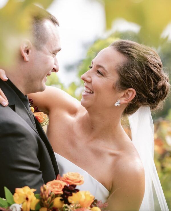 Bride and groom smiling, embracing outdoors. Woman in white dress, man in black suit. Sunlit foliage in background.