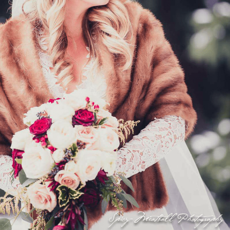 Bride in fur shawl holds bouquet with white and burgundy roses; lace gloves.