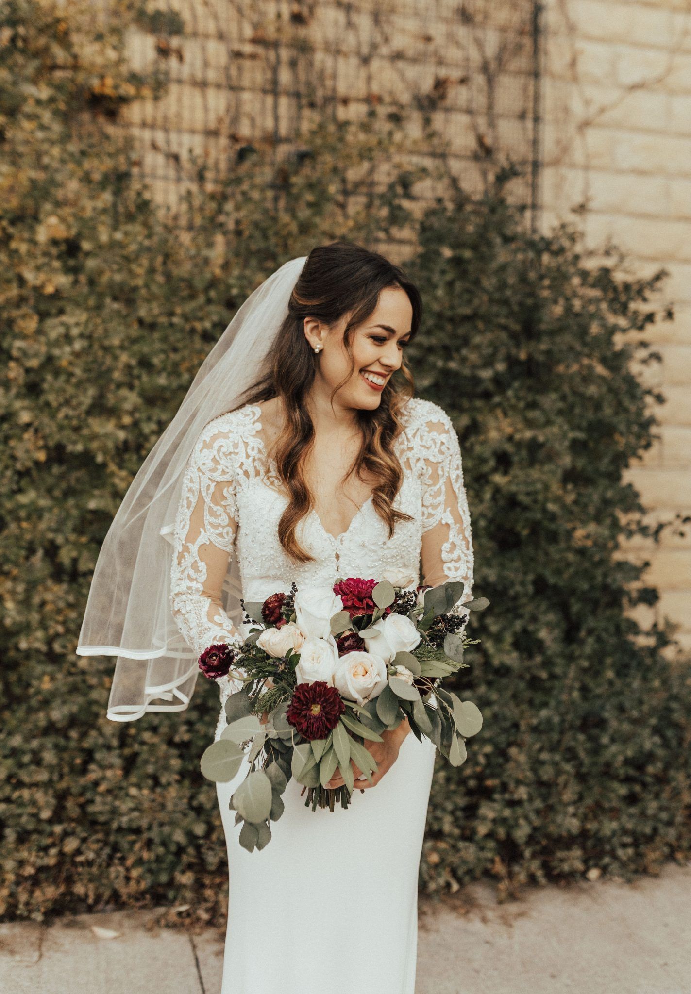 Bride in white lace dress smiles, holding bouquet outdoors.