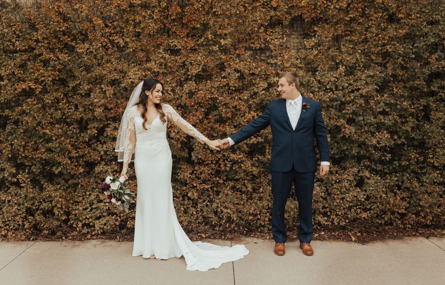 Bride and groom holding hands, looking at each other, standing in front of a brown hedge.