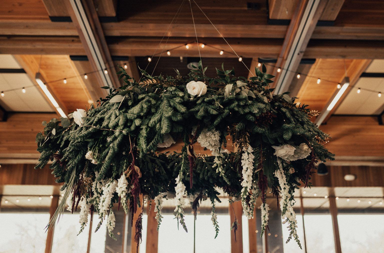 Floral chandelier with greenery and white flowers, hanging from wood beams, with string lights.