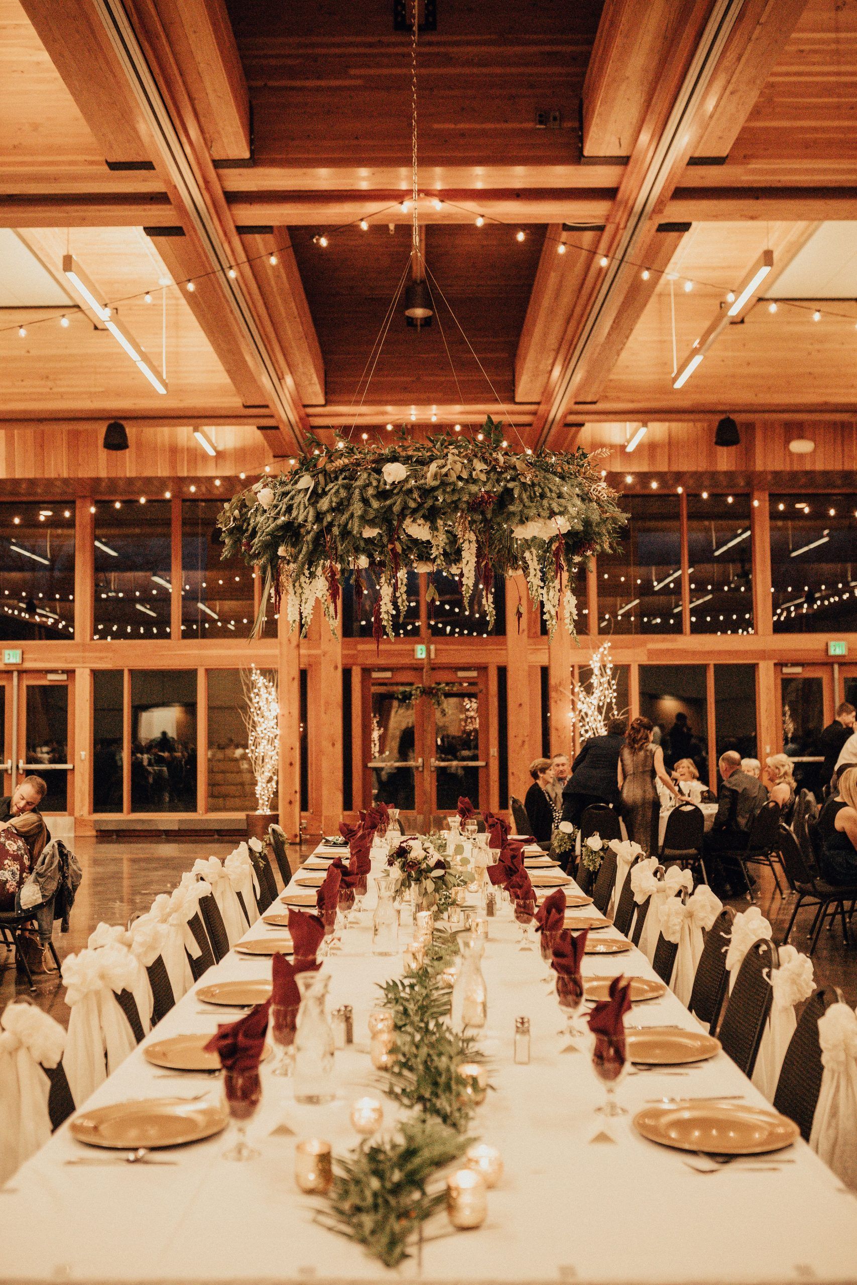 Long dining table set for a wedding reception, centered under floral decor, wood interior, guests seated.