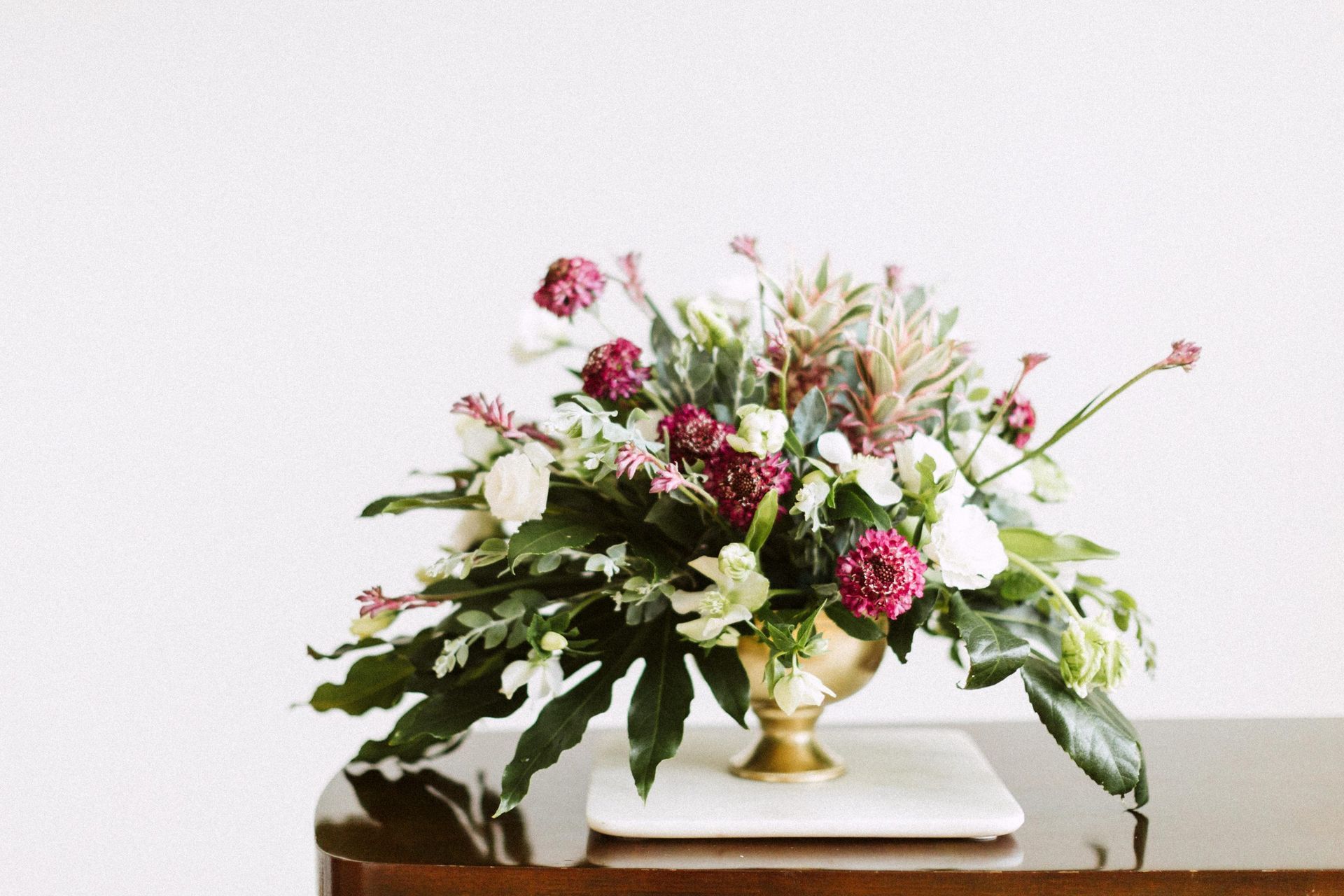 Floral arrangement in gold vase on marble and wooden table, white backdrop.