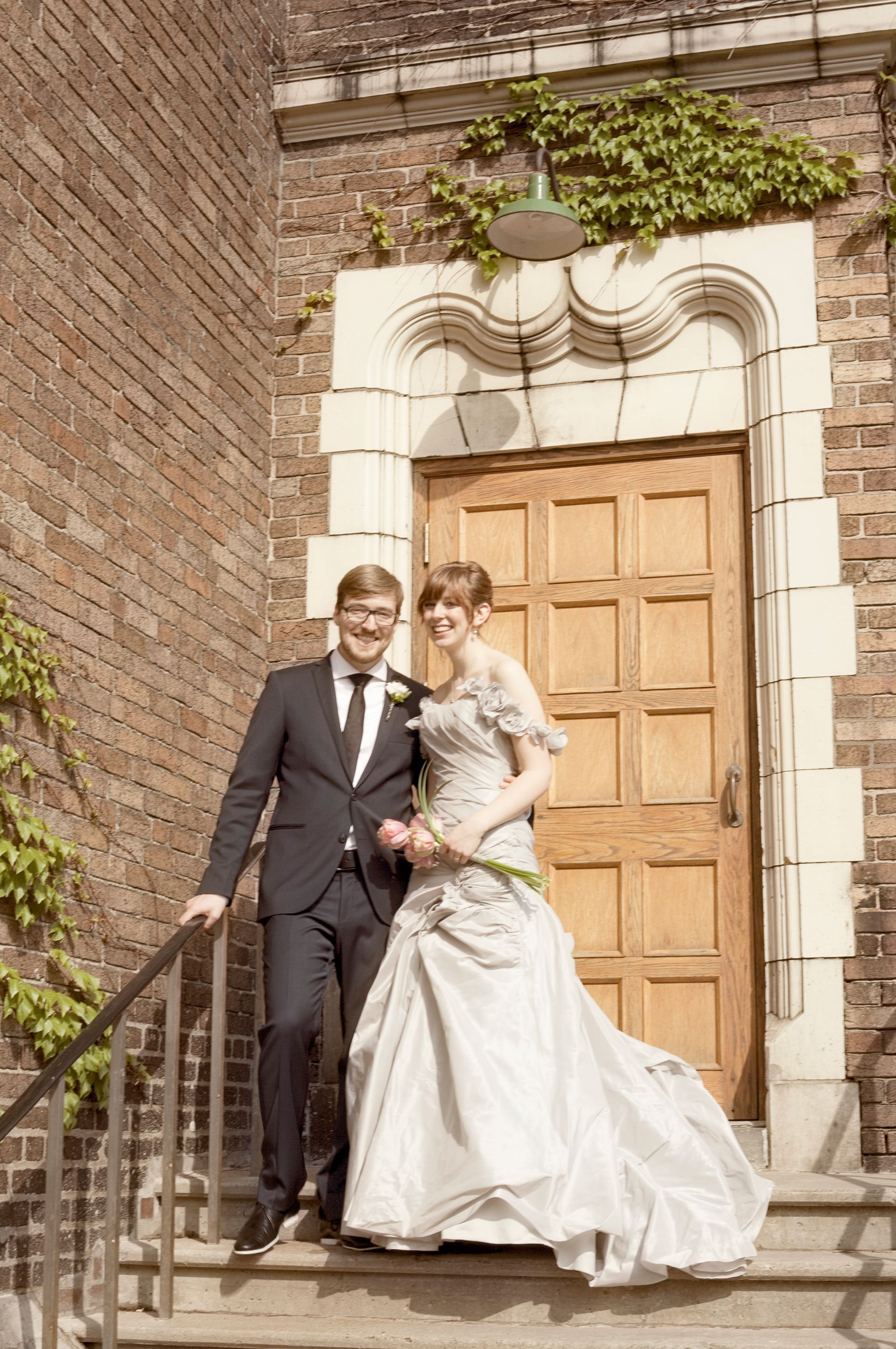 Bride and groom standing on steps in front of a wooden door. Building is brick, with vines growing.
