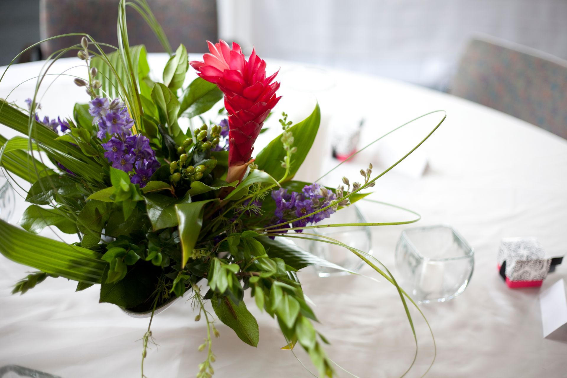 Floral centerpiece with red ginger flower and purple flowers on a white tablecloth.