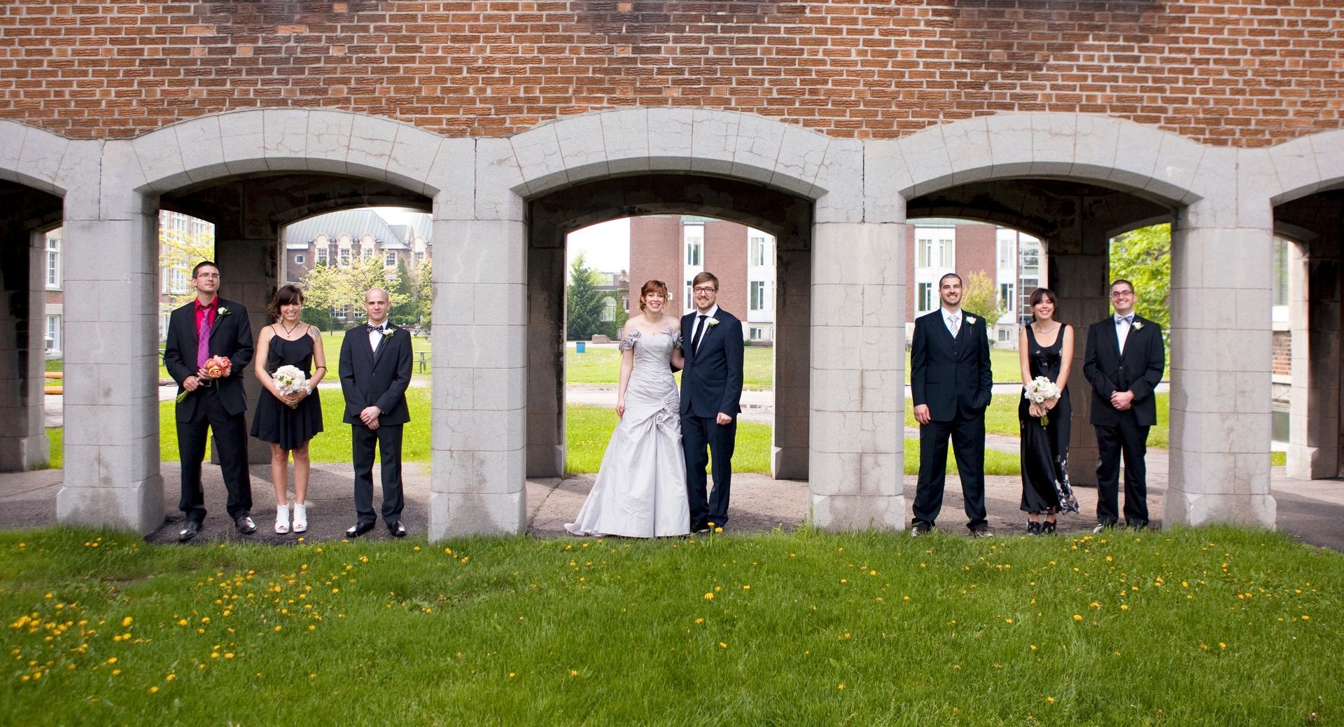 Wedding party posing under brick archways in a grassy area.