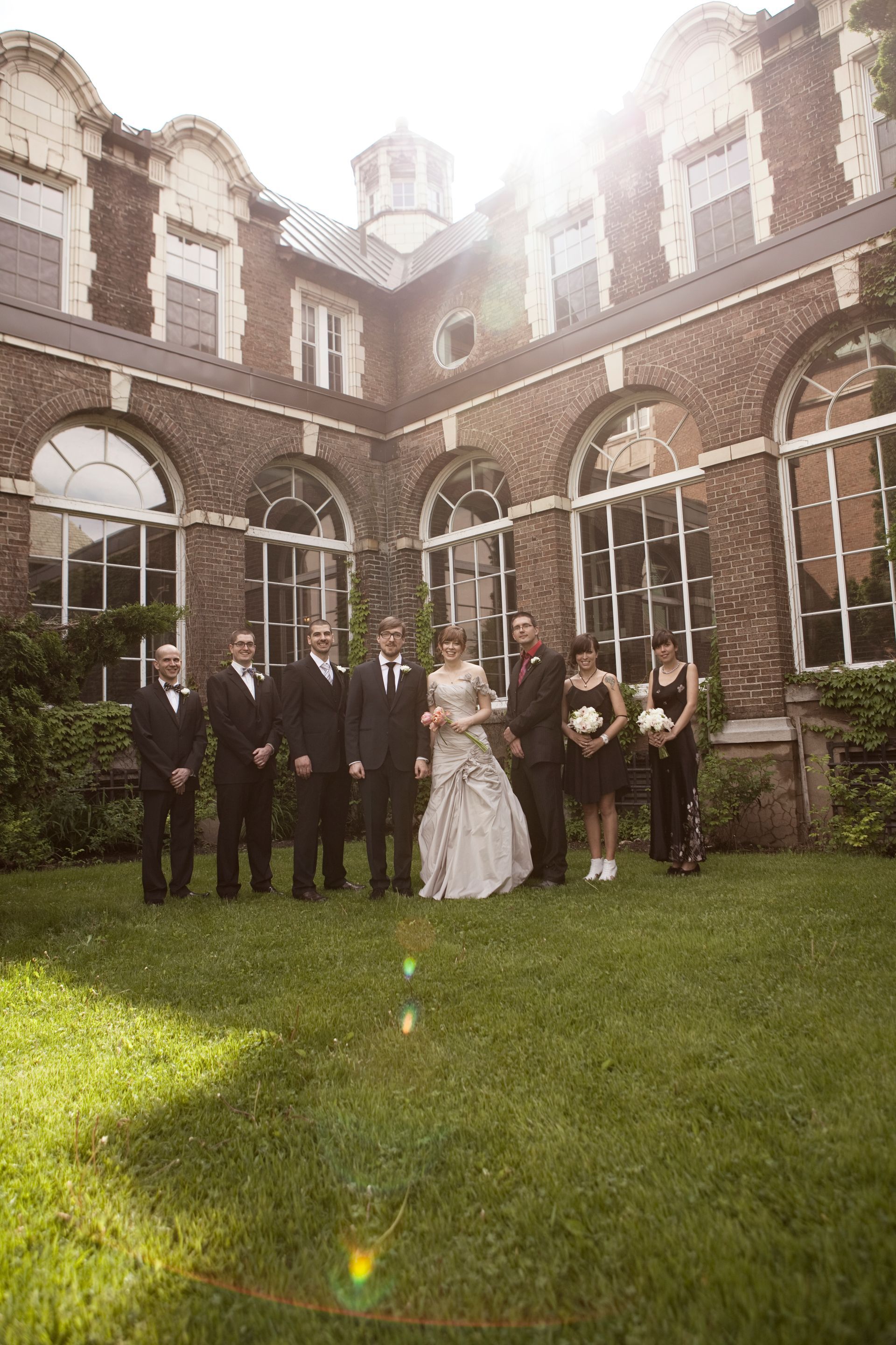 Wedding party poses on the grass in front of a brick building; sunlight shines brightly.