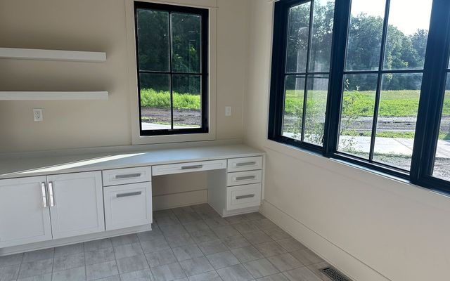 Empty office with white cabinets, desk, and large black-framed windows overlooking greenery.