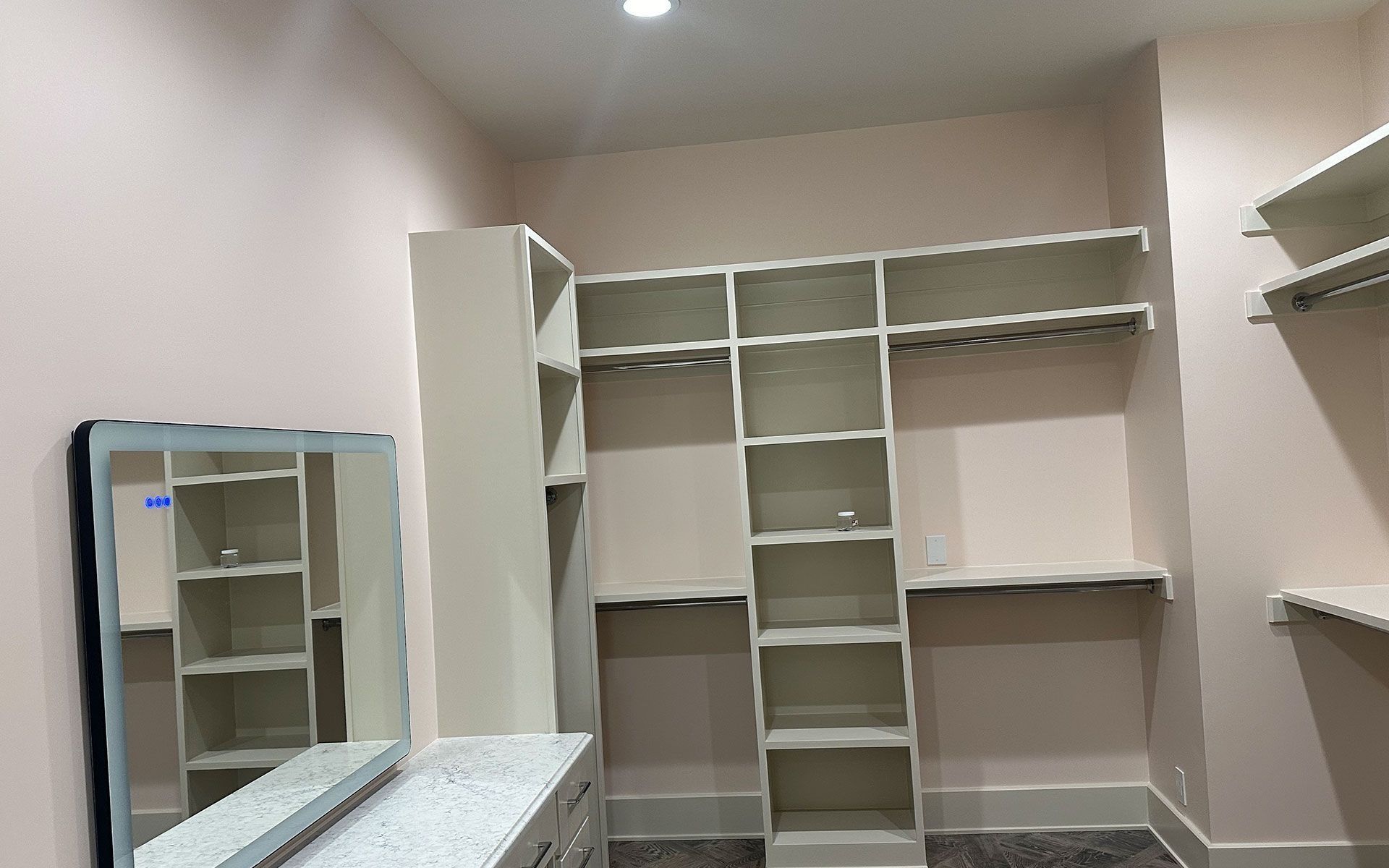 Empty walk-in closet with built-in white shelving and hanging rods, beige walls, and a mirrored dresser.