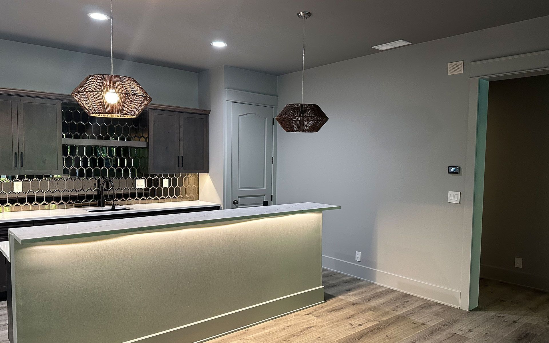 Modern kitchen with gray cabinets, lit island, pendant lights, and dark doorway to the right