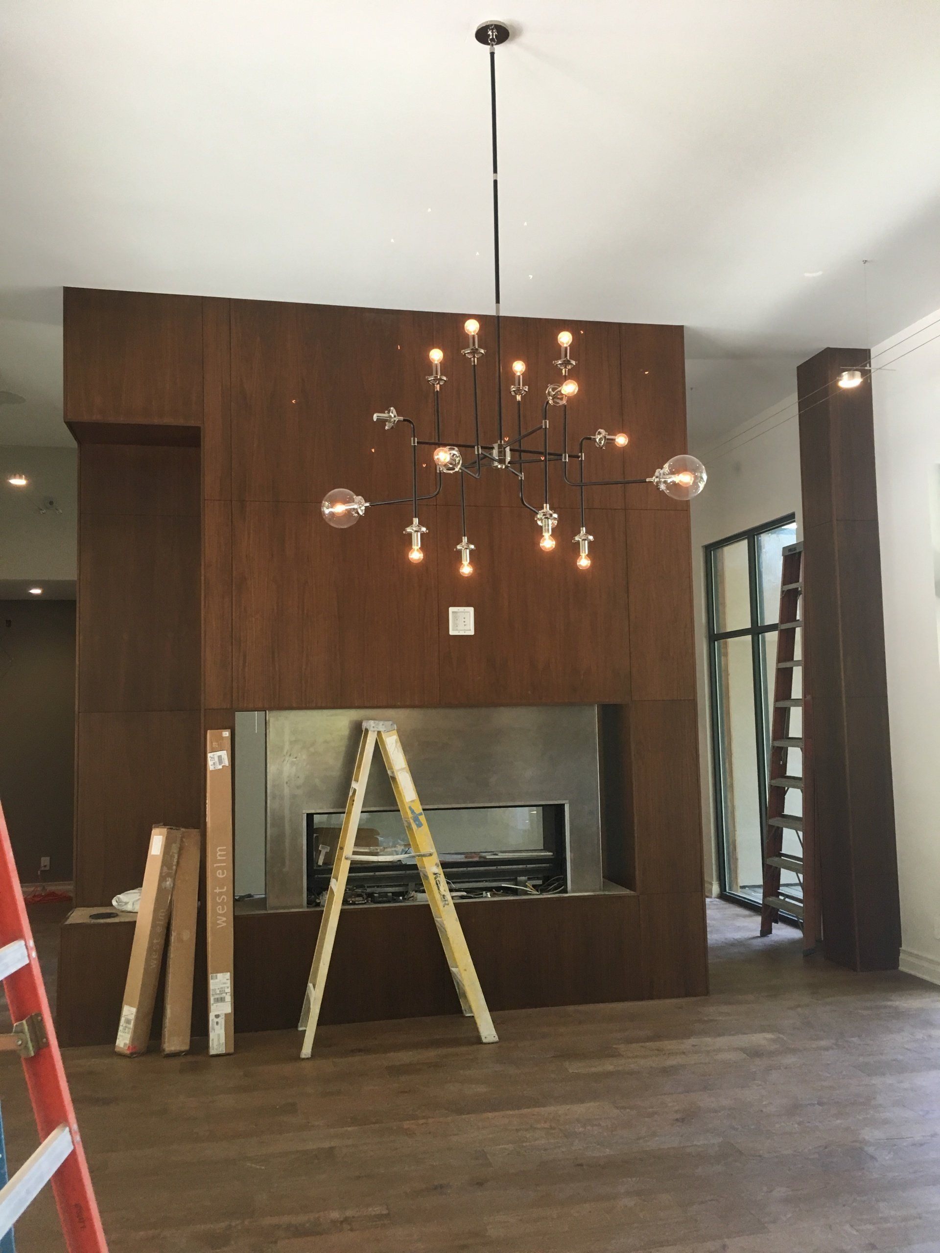 Modern living room with wood-paneled fireplace wall and hanging chandelier under construction