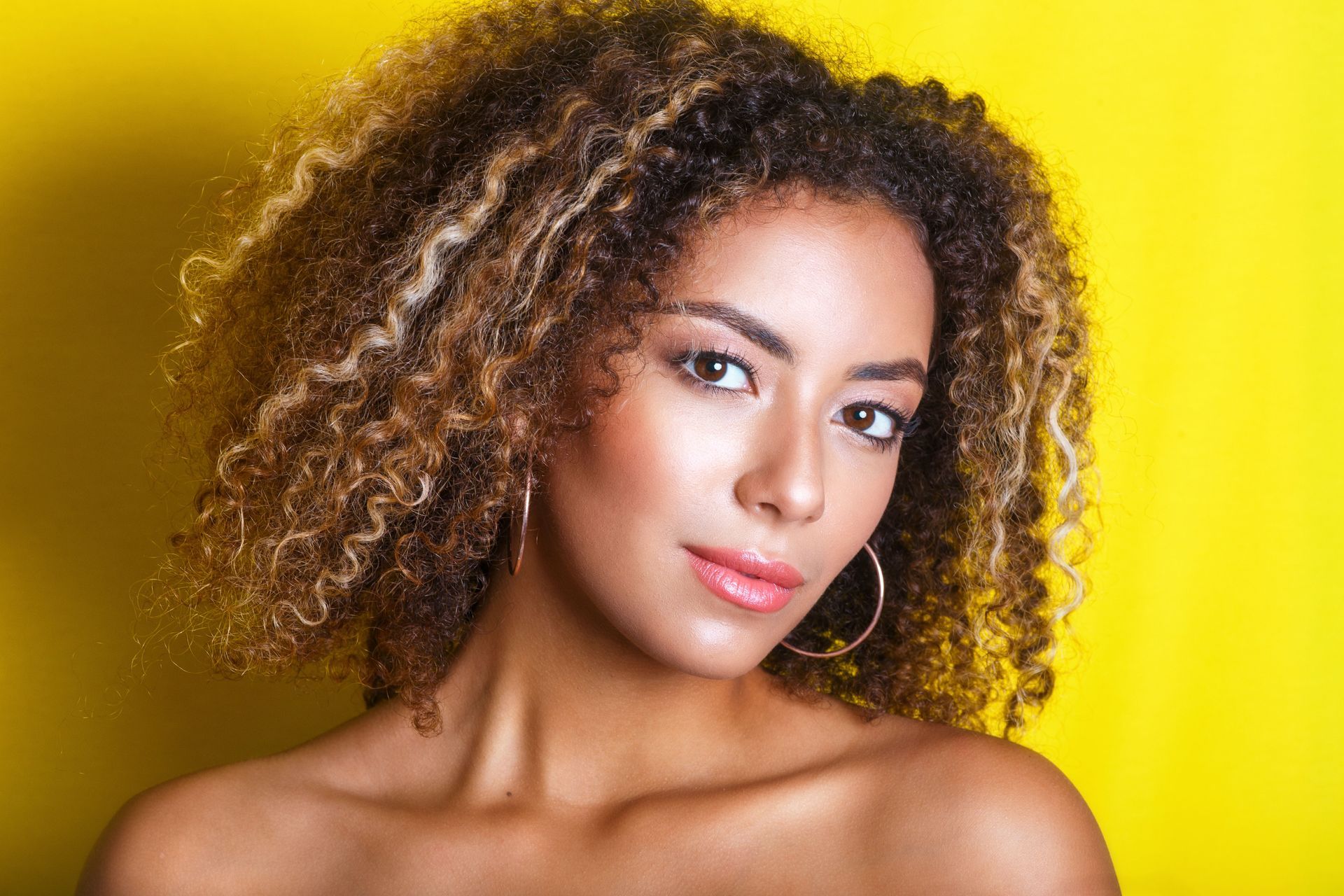 A woman with curly hair and hoop earrings is standing in front of a yellow background.