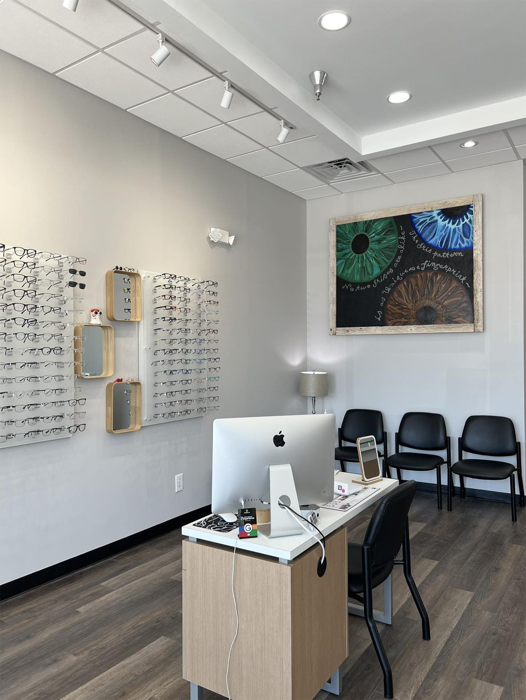 Optical shop interior: desk with computer, eyeglasses display, artwork of eyes, chairs.