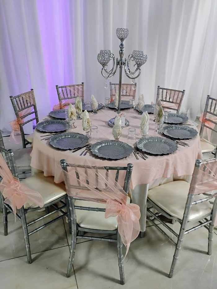 Round table set for a party with pink tablecloth, gray plates, silver chairs, and a candelabra centerpiece.