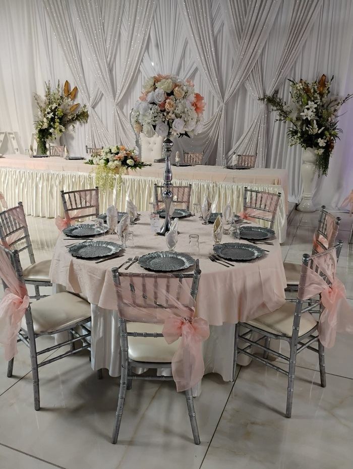 Formal dining setup: round table with silver chairs, pale pink tablecloth, and floral centerpiece.