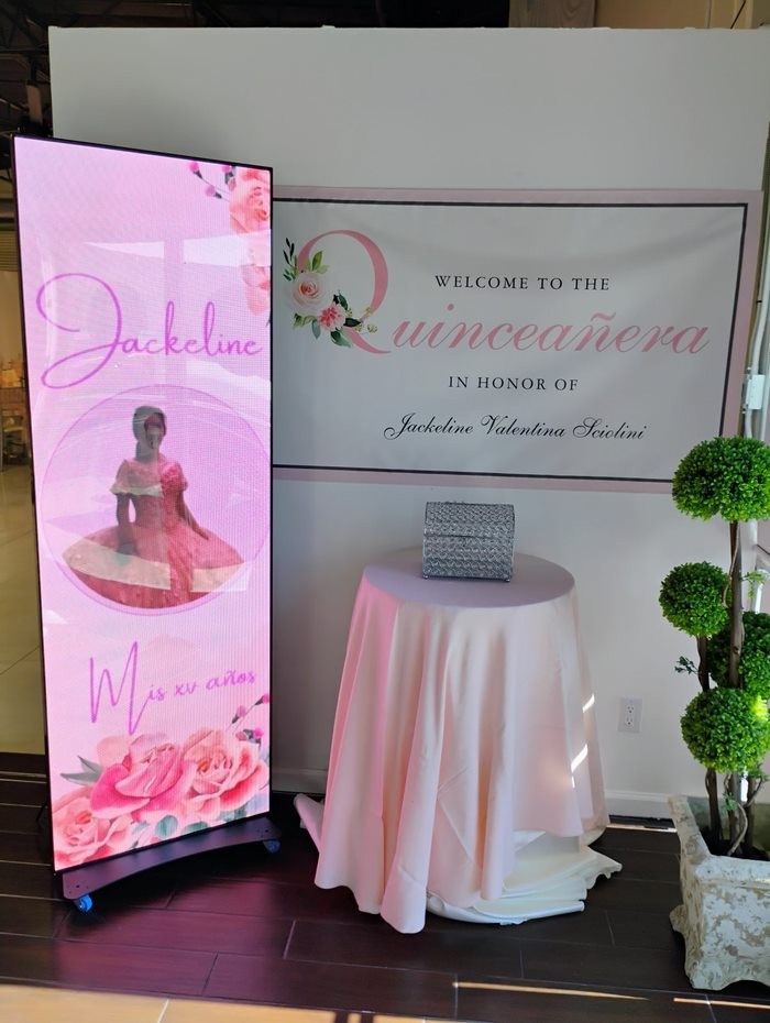 Pink Quinceañera decoration: banner and digital display with girl's photo and event details, round table, and plant.