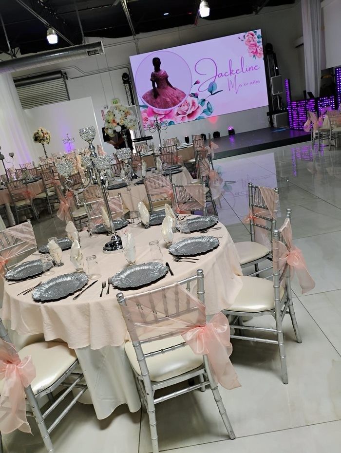 A decorated party venue with round tables set for guests; featuring peach and silver accents, and a screen with a name.