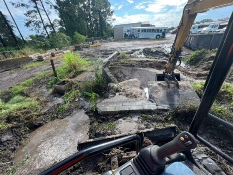 Excavator clearing rubble at a damaged construction site with a building in the background
