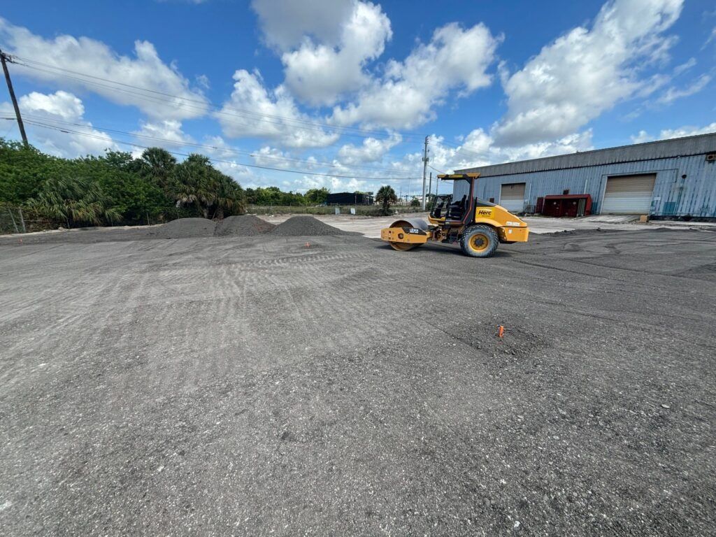 Yellow construction vehicle on a gravel lot beside a warehouse under a blue, cloudy sky