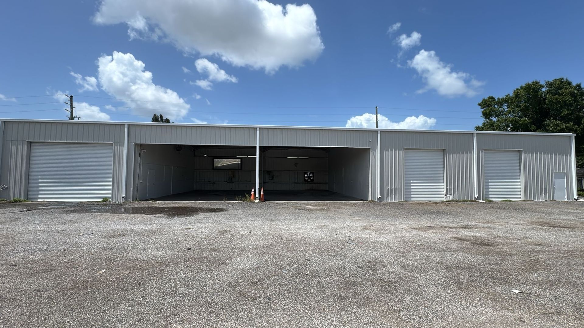 Long metal warehouse with open bays and gravel lot under a blue, cloudy sky