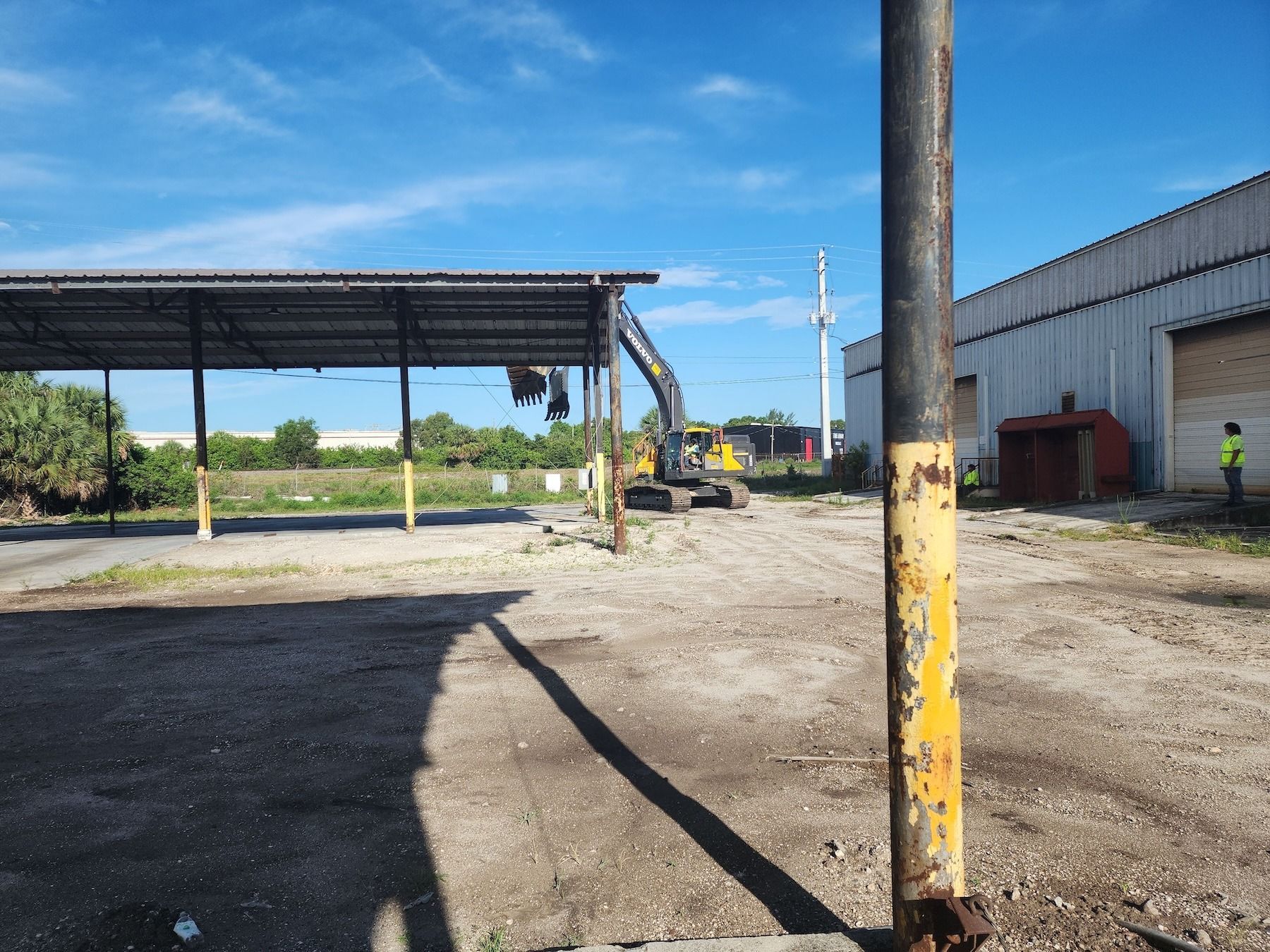 Construction yard with excavator, metal shed, and yellow-black striped pole in the foreground