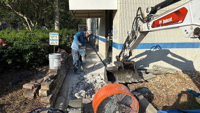 Worker repairs concrete beside building with jackhammer, orange mixer, and tools along a narrow walkway