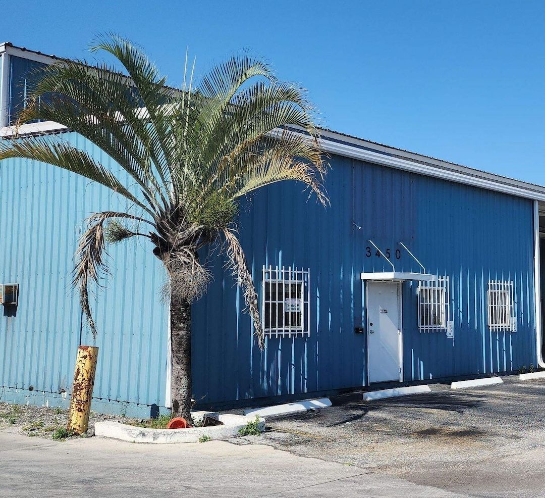 Blue corrugated metal building with white doors and a palm tree in front under a clear sky.