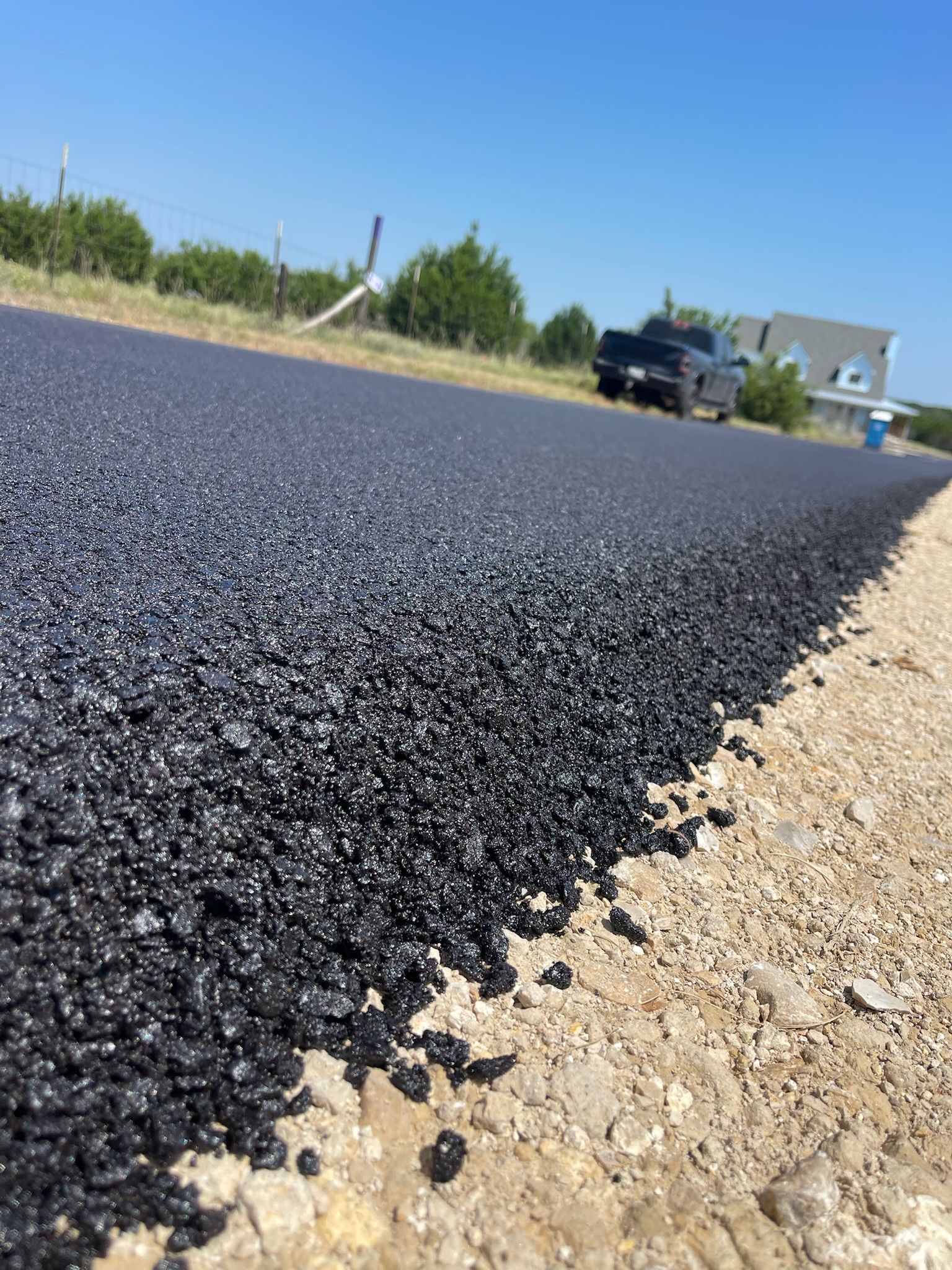 Freshly paved black asphalt road with a gravel shoulder under a blue sky.