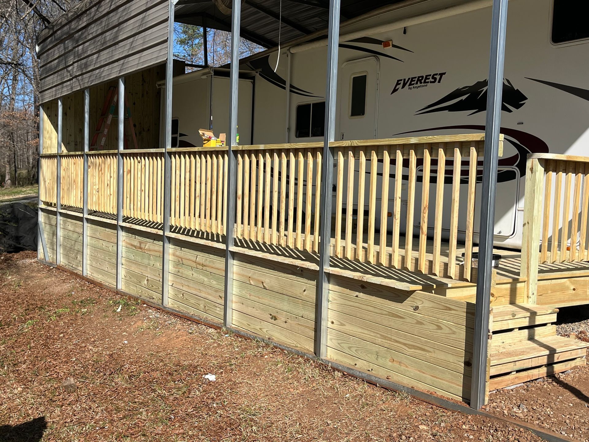 A rv is parked under a covered deck with a wooden railing.