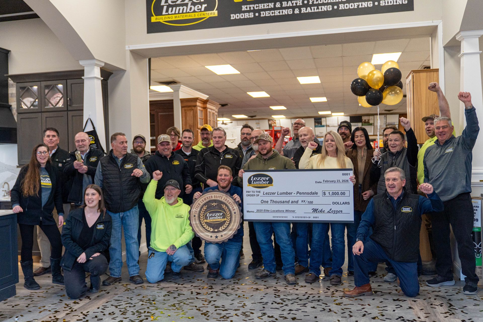 Group of people posing indoors with a large check, trophy, and black-and-gold balloons at a celebration event