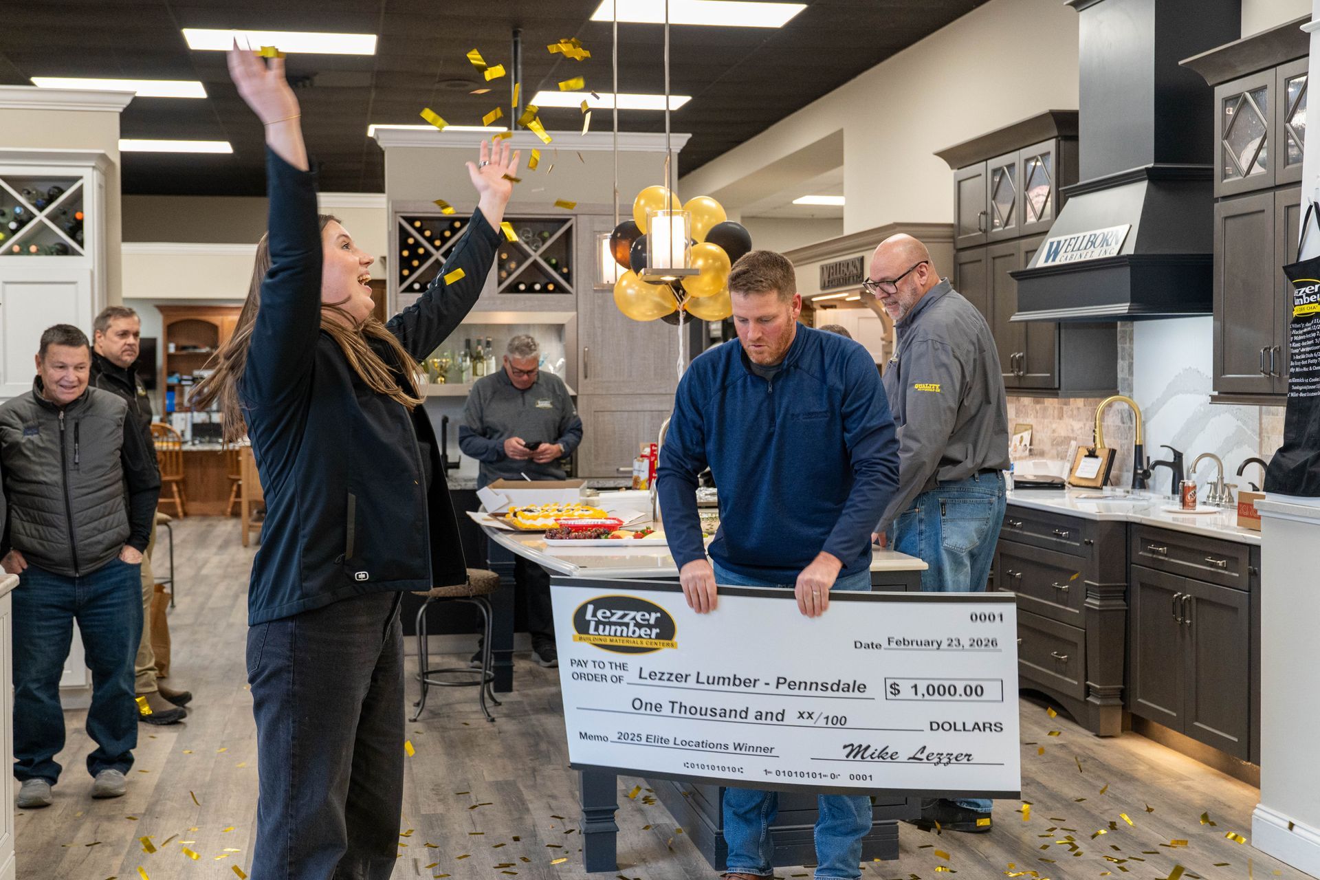 People in a kitchen showroom gather around a man holding a large ceremonial check.