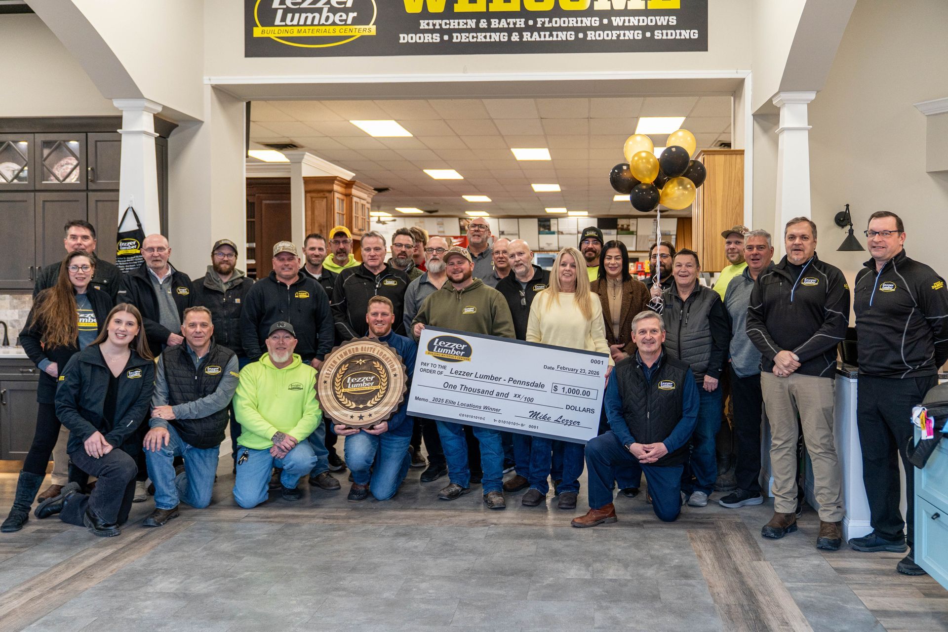Group of people posing inside a storefront, holding a large check and decorative plaque, with black and gold balloons.