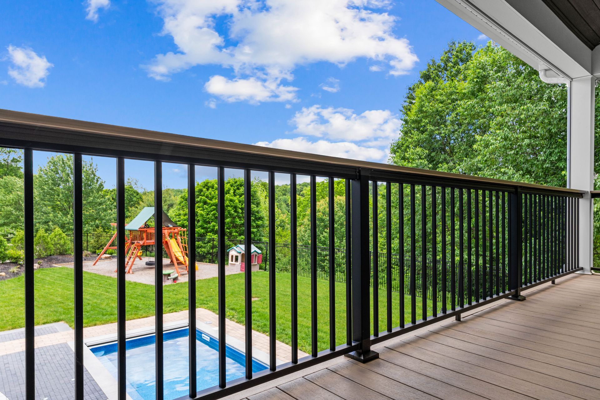 Balcony view over a backyard with black railing, blue pool, green trees, and a playset under a bright sky