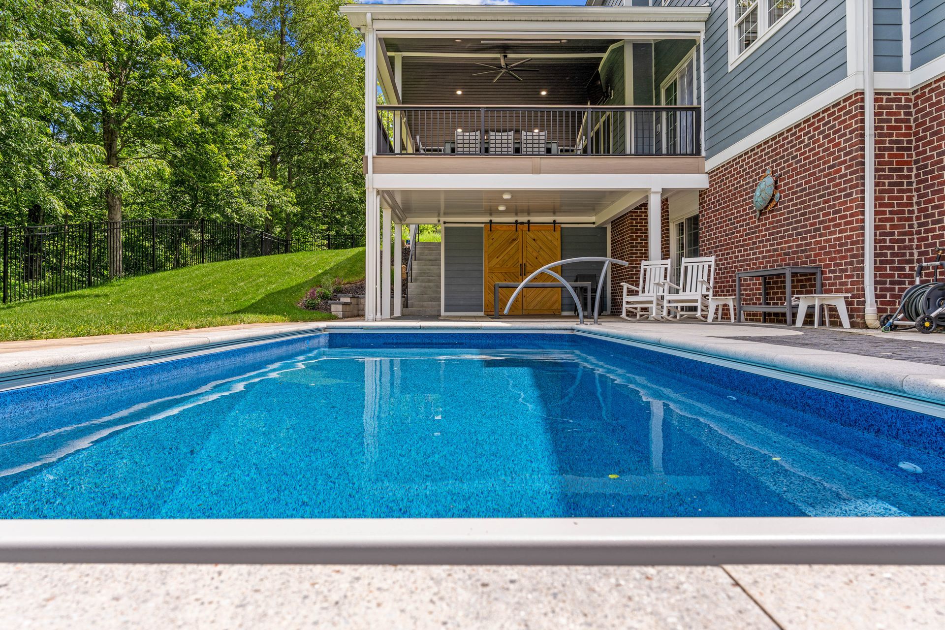 Backyard pool with a house patio, white chairs, and lush green trees beyond