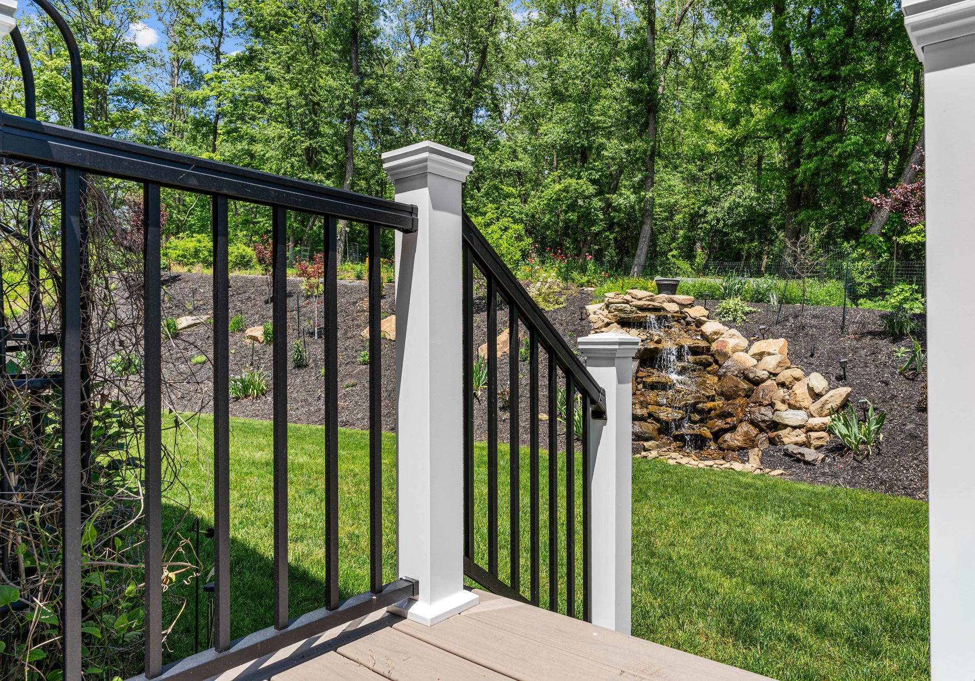 Black metal railing beside a white post on a porch overlooking a grassy yard and stone retaining wall