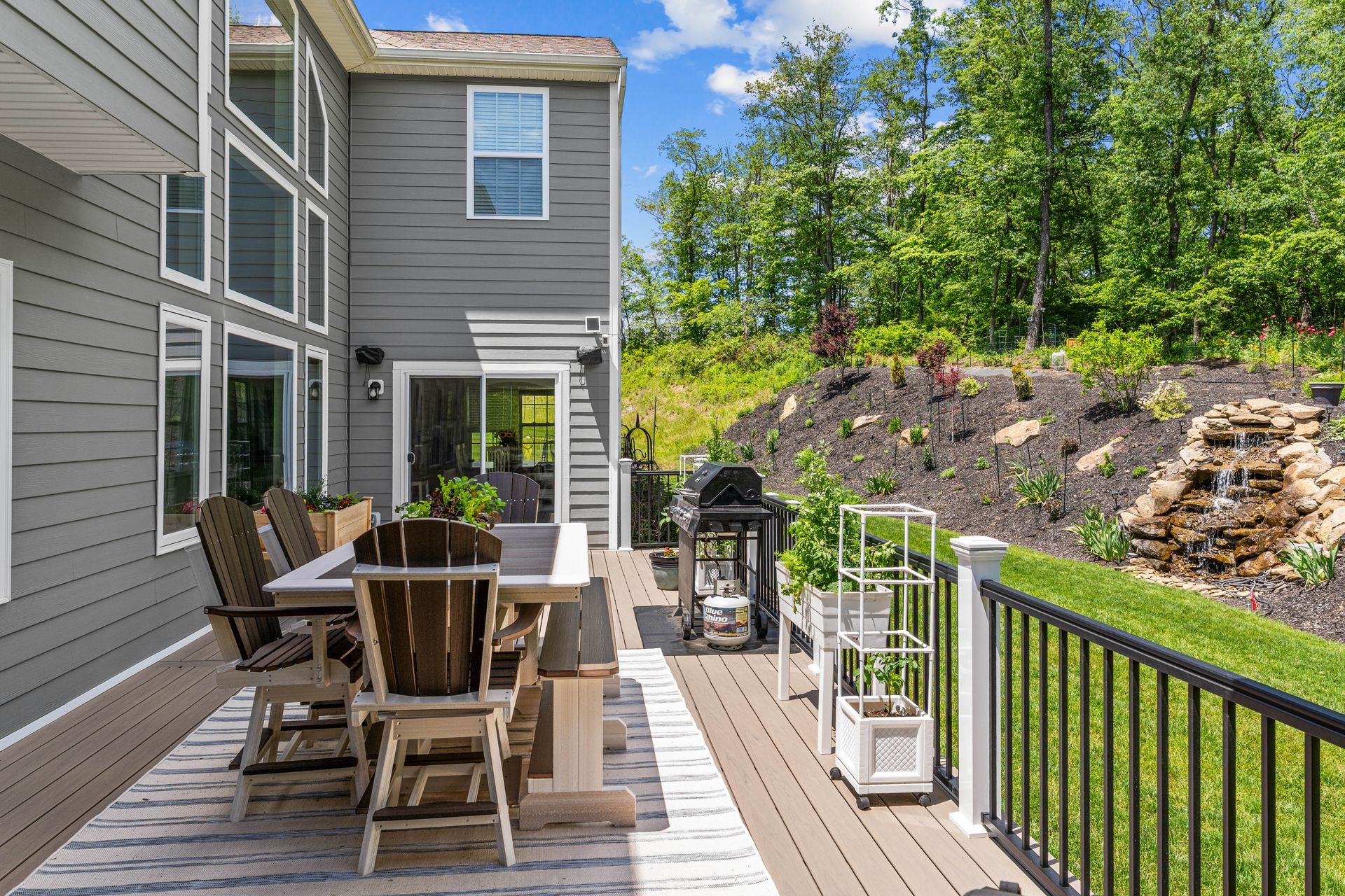 Patio deck with wooden dining set beside gray house, overlooking a wooded hillside and black railing