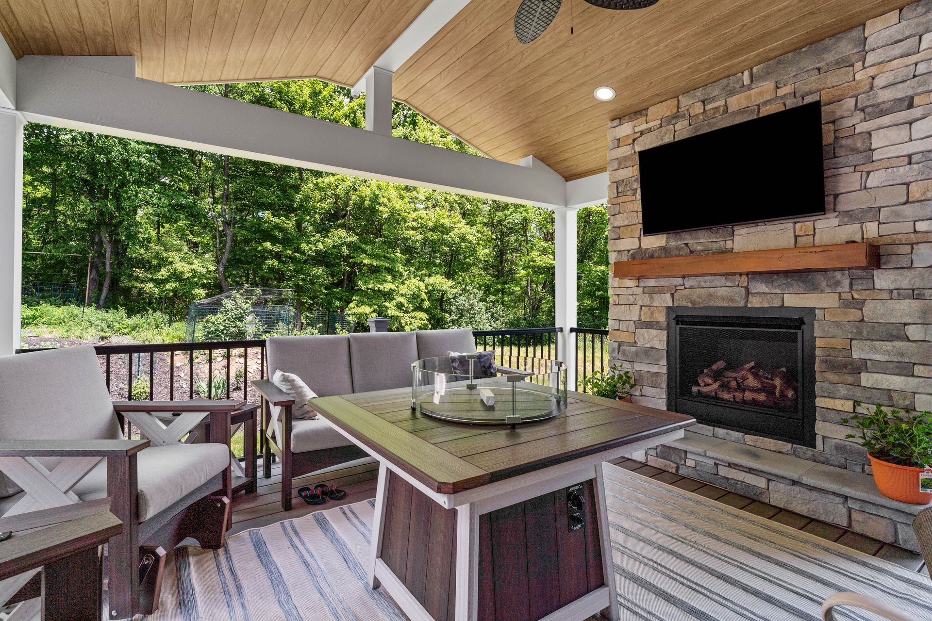 Covered patio with wicker seating, stone fireplace, wall-mounted TV, and green wooded view