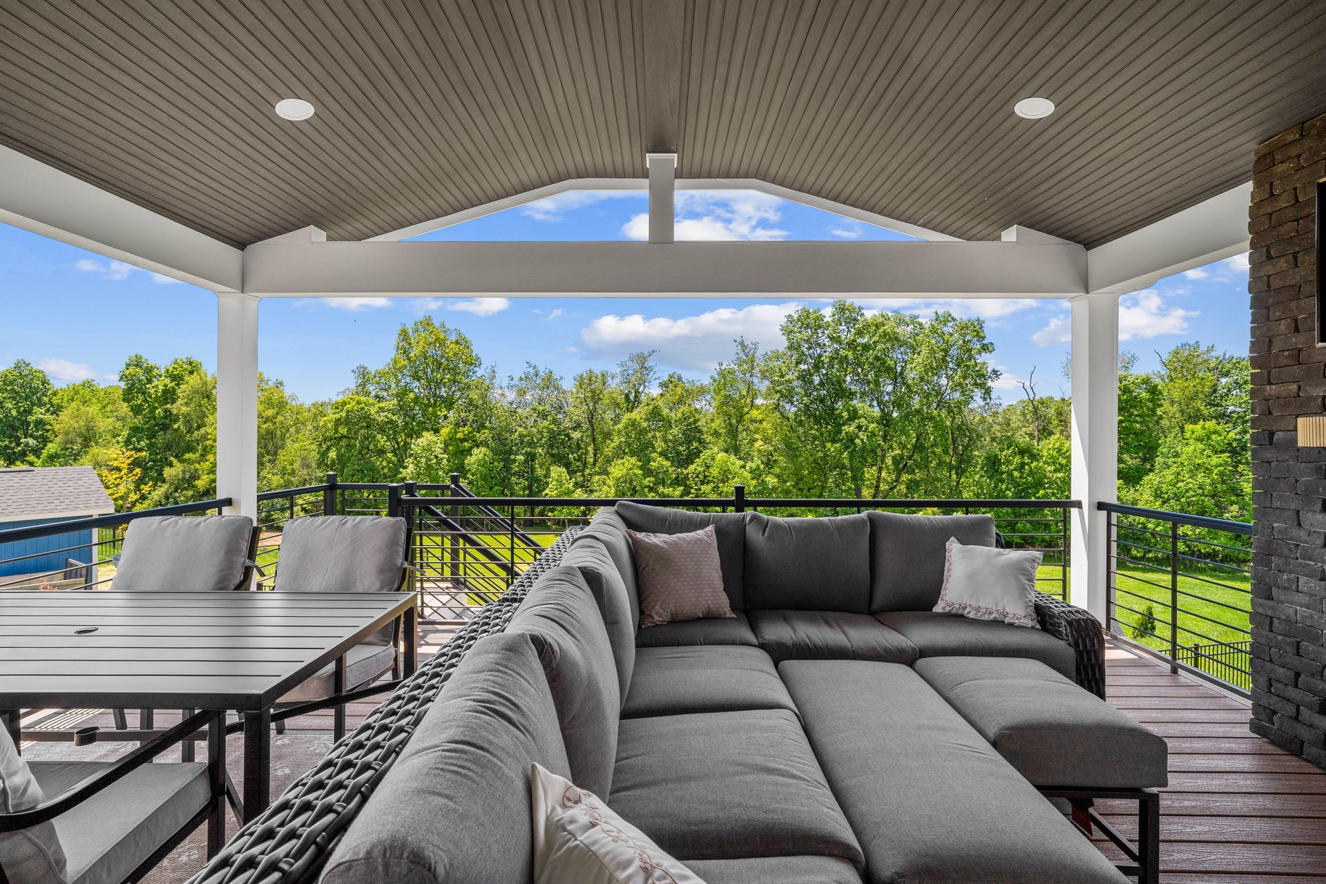 Covered patio with gray sectional seating, dining table, and view of green trees beyond railing