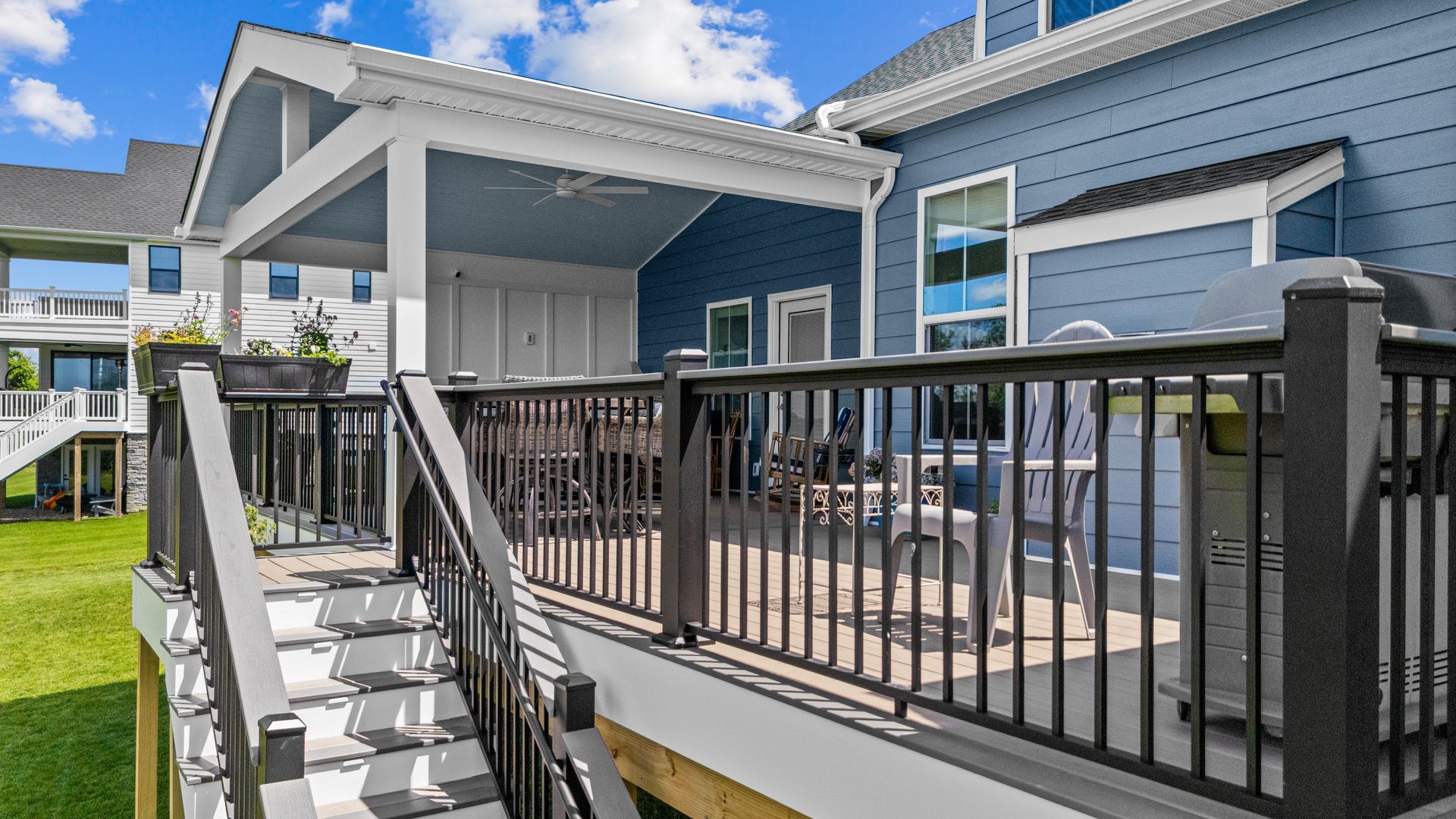 Modern gray house exterior with a raised wooden deck, black railing, and white stairs under a blue sky
