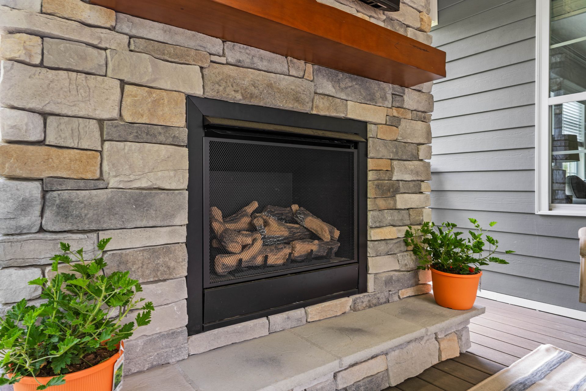 Stone fireplace with a lit gas fire on a covered porch, flanked by potted plants