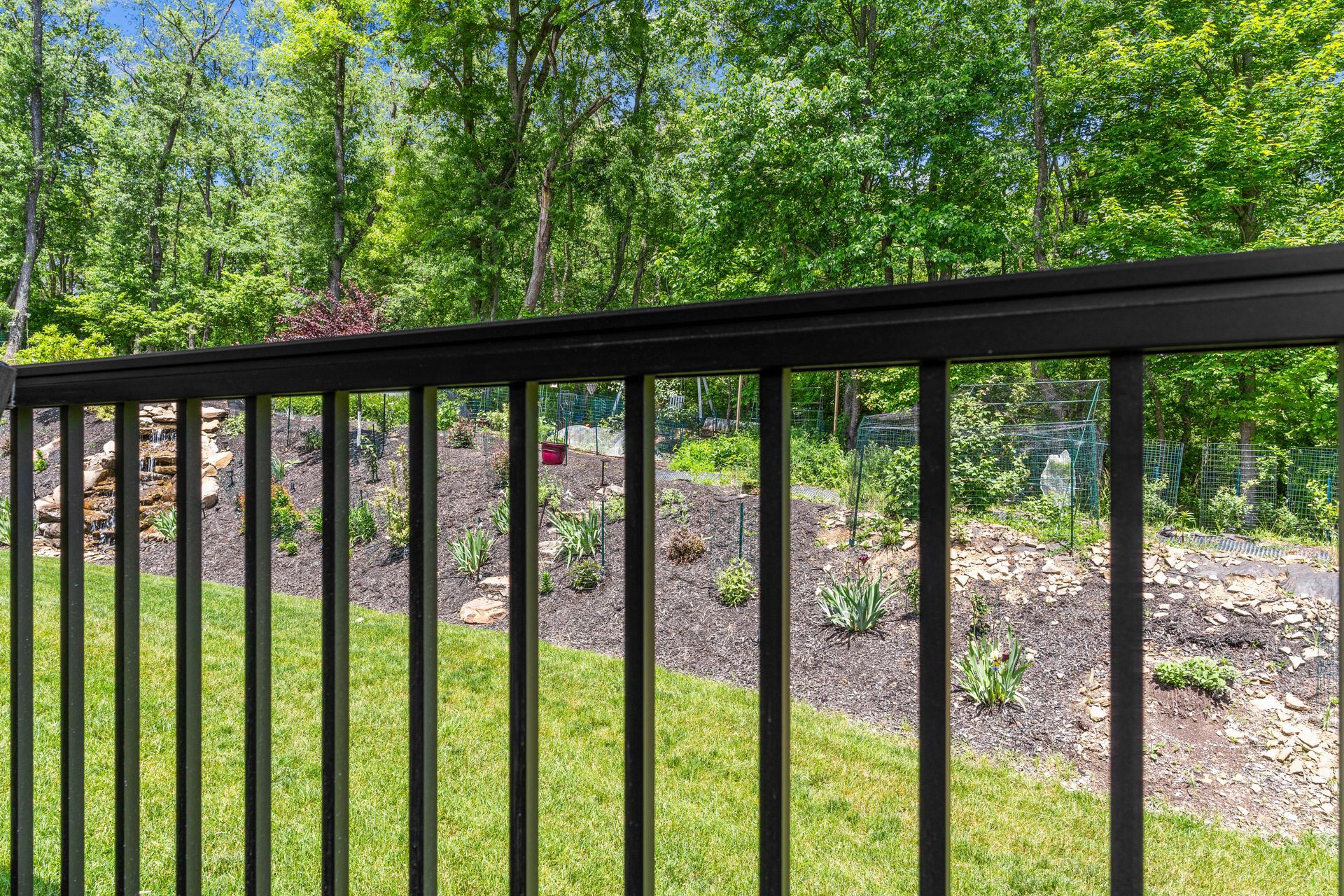 Black metal railing in front of a green yard and wooded hillside with trees and rocks.