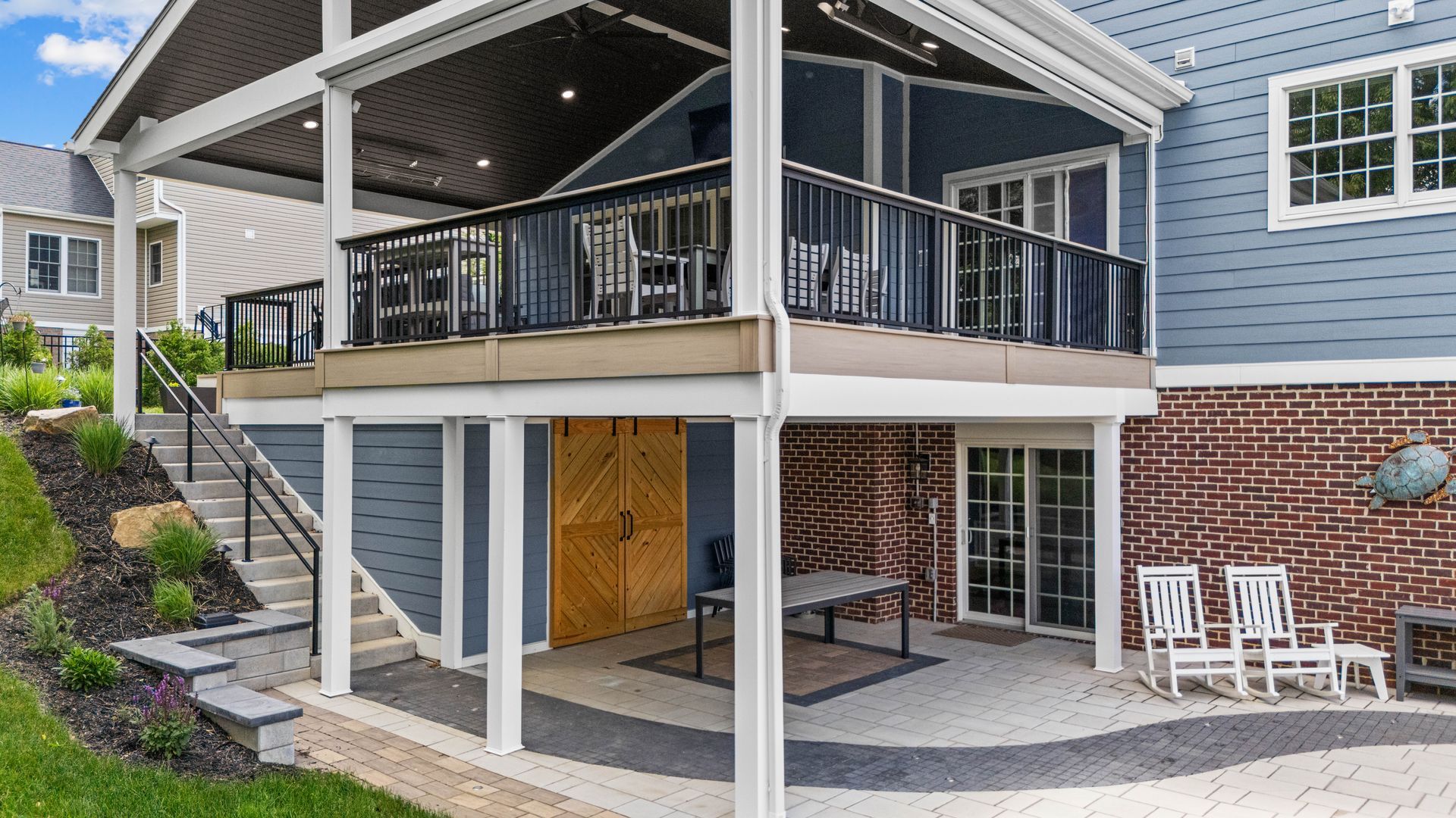 Covered patio under a raised deck beside a brick house with stairs, railing, and wooden doors
