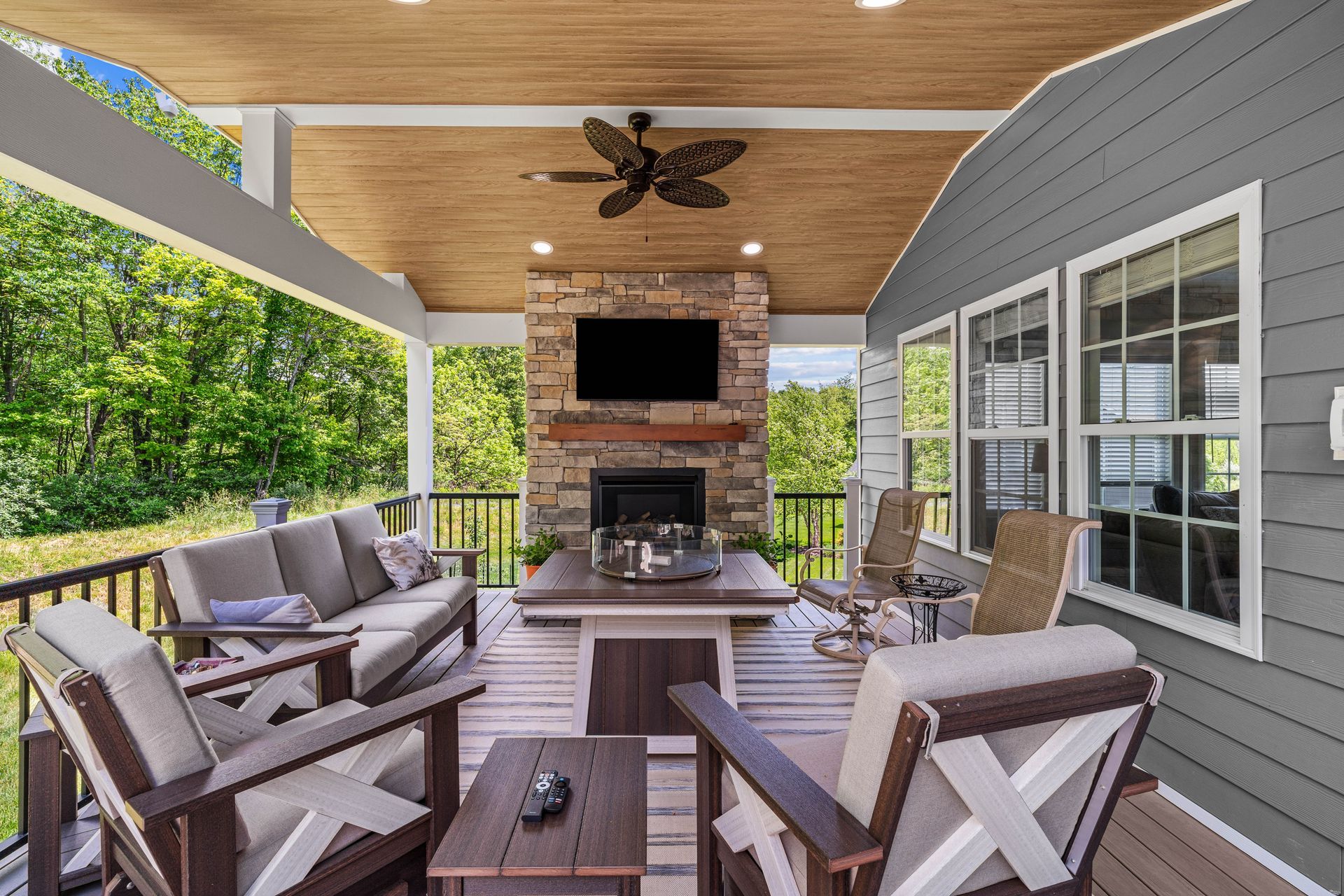 Covered outdoor patio with seating, fireplace, TV, and ceiling fan overlooking green trees