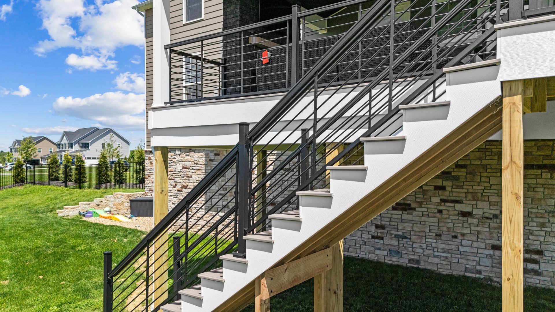 Exterior staircase with black railings beside a stone-and-siding house, overlooking a green yard